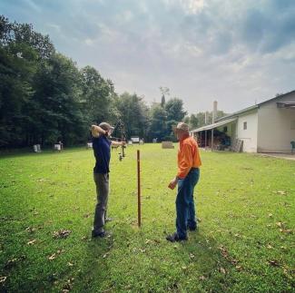 A person stands in a green field and aims a bow and arrow at a target while an instructor looks on
