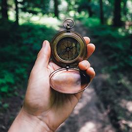A hand holds out a bronze compass with a forest trail in the distance