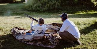 An instructor sits behind a youth hunter as they aim a rifle to the sky during a mentored youth waterfowl hunt