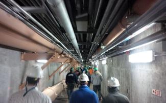 A group of wastewater operators walk in a brightly lit underground tunnel with pipes overhead