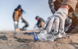 A hand picks up a water bottle discarded on a beach with other volunteers in the background