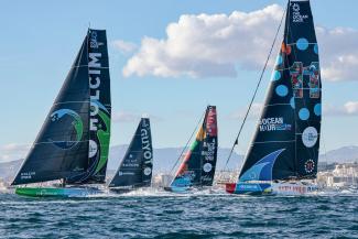 Four high-tech sailing boats with blue sails with a blue sky with puffy white clouds and mountains in the background. Photo credit: Sailing Energy / The Ocean Race