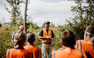 A DEM Biologists wearing an orange vest speaks to a group of people, also wearing orange vests, at a newly created young forest area at the Great Swamp Management Area in West Kingston