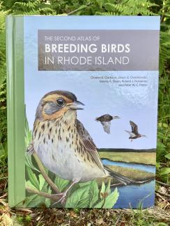 A hardcover book with pea green spine titled Breeding Birds in Rhode Island with a large songbird pictured on the cover sits among a bright green, forested landscape