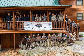 A group of hunters standing on the deck of a log building with others holding wild turkeys they harvested as part of a mentored hunt.