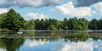 Evergreen trees are reflected in a freshwater pond with two anglers in a canoe at Wickaboxet Management Area in West Greenwich