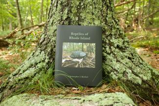 The Reptiles of Rhode Island book with a black cover and image of a painted turtle. The book is leaning up against a moss covered tree with plants and brown leafy debris underneath. 