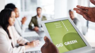 Hands holding a tablet with the words "Go Green" with an office setting of people seated at a conference table in the background