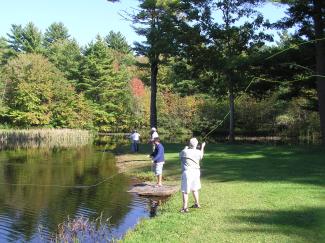 A photo shows three people fly fishing on the shoreline of a pond. 