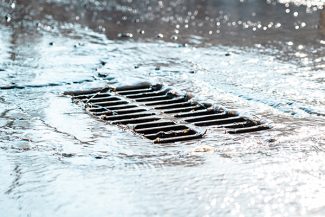 The grate of the storm sewer after the rain. The water drains into the storm drain. Sun glare, defocused background