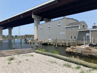 A gray two story building surrounded by the Sakonnet River with the bridge overhead