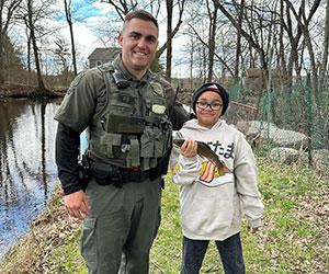 RI Environmental Police Officer smiles alongside a youth angler holding a rainbow trout on the opening day of freshwater fishing season 